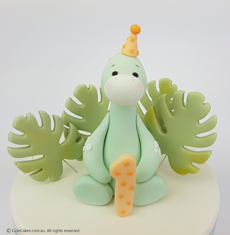 May include: A light green dinosaur cake topper with a party hat and the number one. The dinosaur is surrounded by green leaf decorations. The cake topper is on a white and light blue cake.