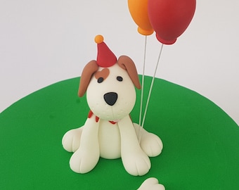 May include: A white fondant dog wearing a red and yellow party hat sits on a green fondant cake. The dog has brown ears and a red collar. Two balloons, one orange and one red, are attached to the cake behind the dog.