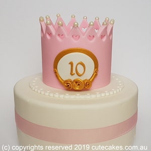 Puede incluir: Un pastel de dos pisos decorado con glaseado rosa y blanco. El piso superior tiene una corona rosa con detalles de perlas blancas y un óvalo dorado con el número "10" en su interior. El piso inferior tiene una cinta rosa y glaseado blanco con un borde de perlas blancas.