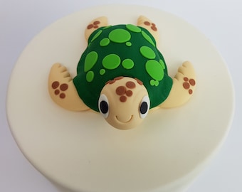 May include: A green and brown fondant turtle cake topper with a smiling face and green spots on its shell.