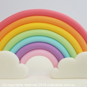Puede incluir: Un adorno de pastel de arcoíris pastel con nubes blancas a cada lado. El arcoíris está hecho de fondant rosa, naranja, amarillo, verde, azul, morado y rosa.