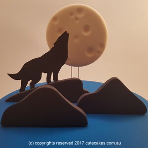 Könnte beinhalten: Kuchendekoration mit einer schwarzen Silhouette eines heulenden Wolfs unter einem Vollmond. Der Mond ist hellbeige mit Kratern. Der Wolf steht auf einer dunkelbraunen Bergkette vor blauem Hintergrund.