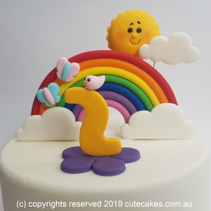 Puede incluir: Un pastel blanco decorado con un arcoíris, nubes, un sol, mariposas y un número dos amarillo. El pastel es perfecto para un segundo cumpleaños.