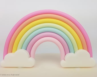 May include: A colorful rainbow cake topper with pastel shades of pink, orange, yellow, green, and blue. Two white cloud accents are at the base of the rainbow. The cake topper is a decorative element for a cake.