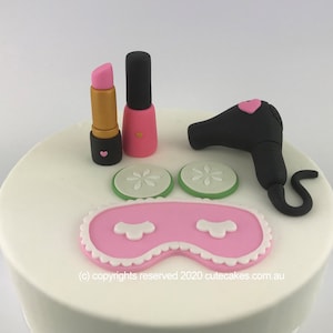 May include: A white cake decorated with pink and black fondant. The cake features a pink eye mask, two green cucumber slices, a pink lipstick, a black nail polish bottle, and a black hair dryer with a pink heart.