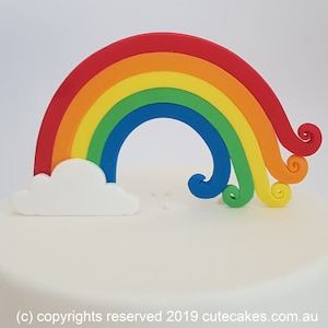 May include: A white fondant cake with a rainbow and cloud topper. The rainbow is made of red, orange, yellow, green, blue, and purple fondant. The cloud is white fondant.