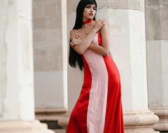 May include: A woman with long black hair wearing a red and pink dress stands in front of a white pillar. She is looking down and has her arms crossed.