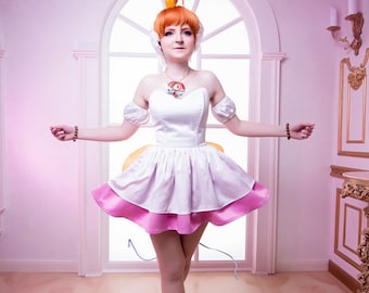 May include: A woman in a white and pink dress with a heart-shaped pendant necklace and a crown on her head. She is standing in a room with a large arched window and pink walls.