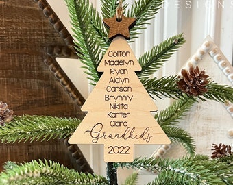 May include: A wooden Christmas tree ornament with the names Colton, Madelyn, Ryan, Aydyn, Carson, Brynny, Nikta, Karter, Clara, and the text "Grandkids 2022".