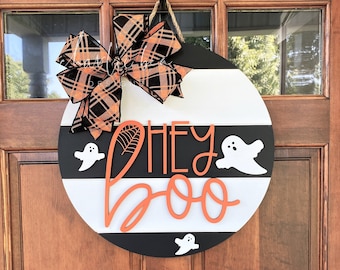 May include: A black and white striped round door hanger with the text "Hey Boo" in orange. The door hanger has a black and orange plaid bow and three white ghost silhouettes.