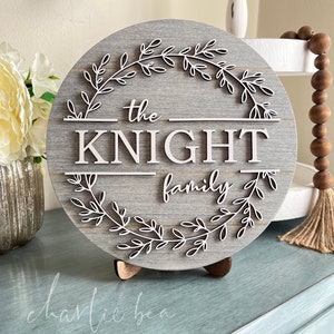 Puede incluir: Un letrero redondo de madera gris con una corona de hojas blancas y el texto "the KNIGHT family" en letras blancas.