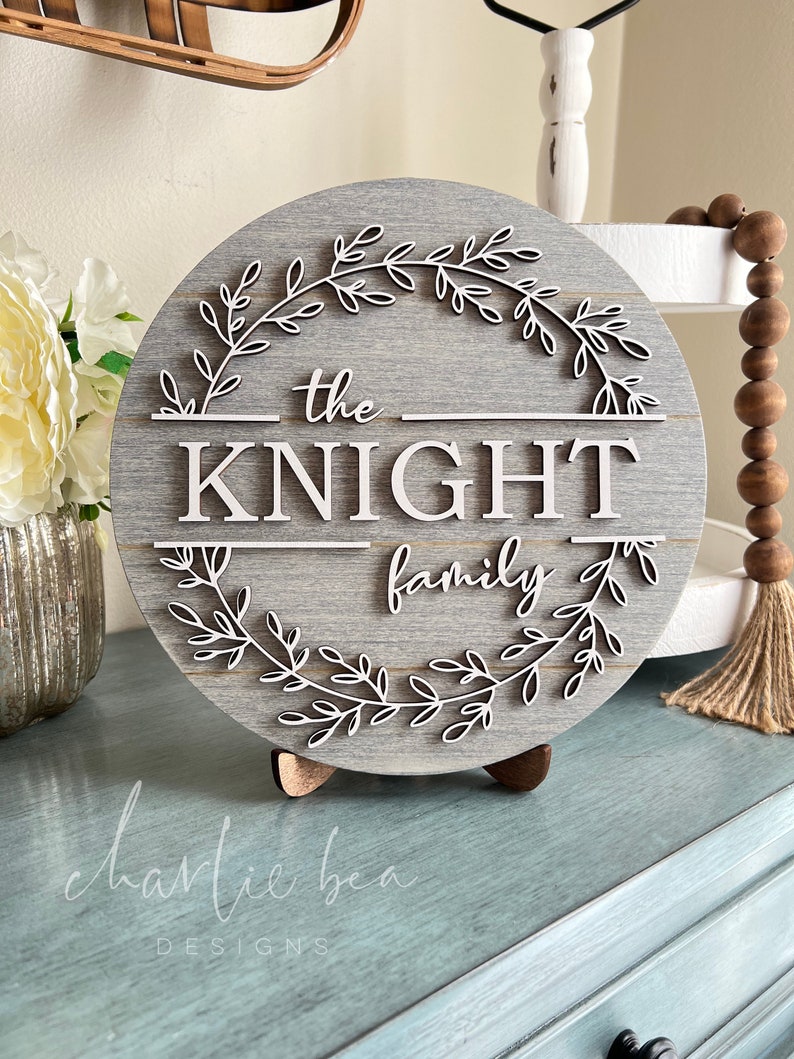 Puede incluir: Un letrero redondo de madera gris con una corona de hojas blancas y el texto "the KNIGHT family" en letras blancas.