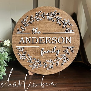 Puede incluir: Un letrero redondo de madera con un dise&ntilde;o de corona blanca y el texto "the ANDERSON family" en letras blancas.