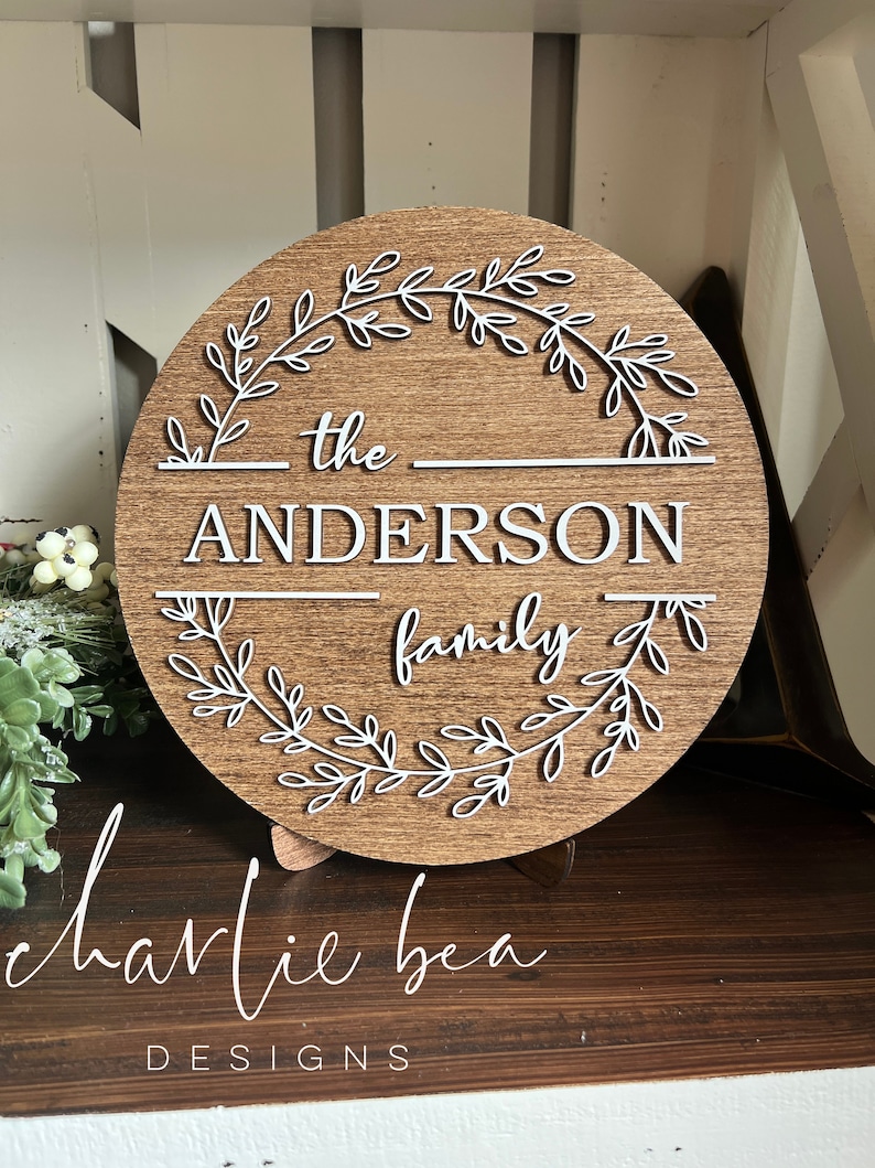 Puede incluir: Un letrero redondo de madera con un dise&ntilde;o de corona blanca y el texto "the ANDERSON family" en letras blancas.
