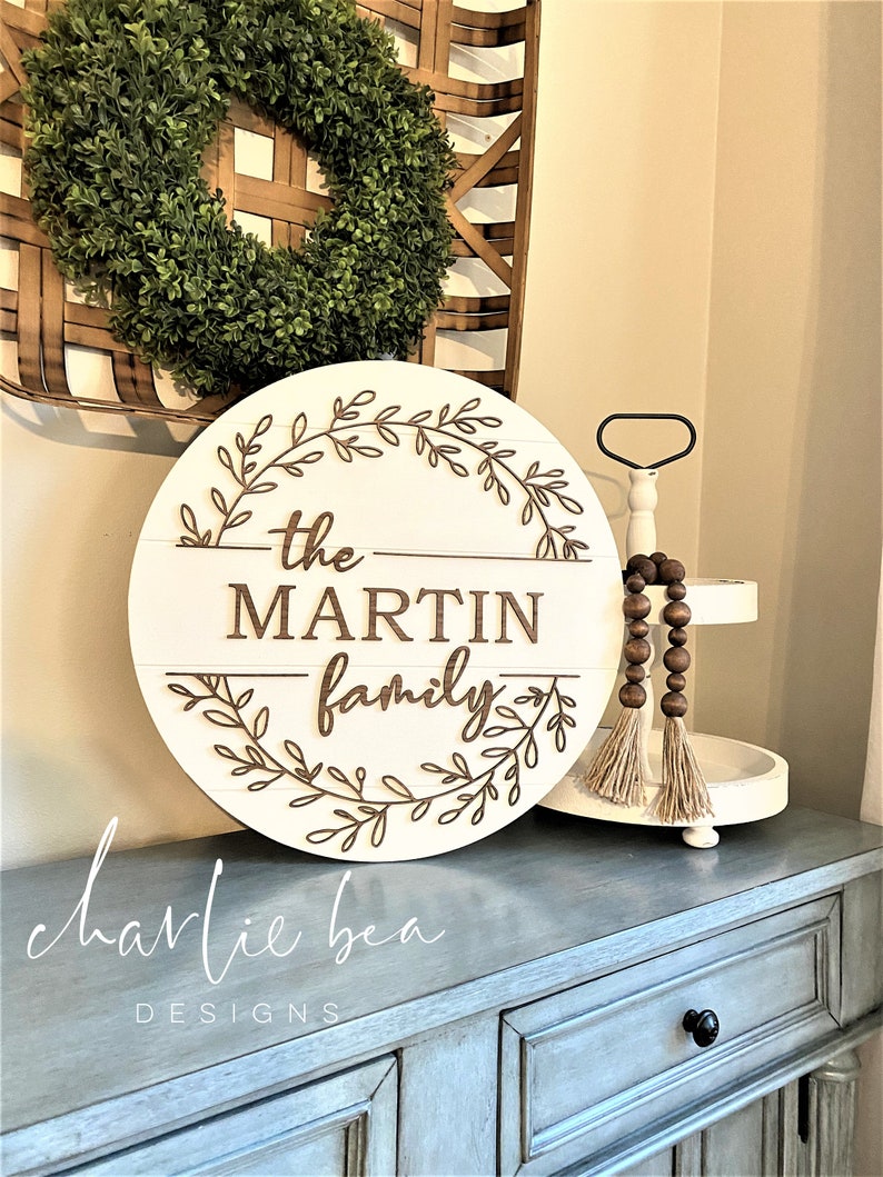 Puede incluir: Un letrero redondo de madera blanca con el texto "the MARTIN family" en letras marrones. El letrero est&aacute; decorado con una corona de hojas marrones. El letrero est&aacute; colocado sobre una c&oacute;moda gris con una bandeja de niveles blanca y una guirnalda de cuentas de madera.