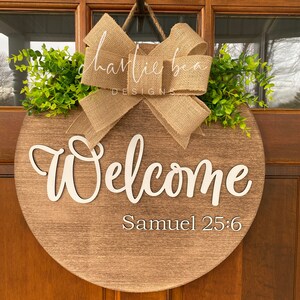 K&ouml;nnte beinhalten: Ein rundes, h&ouml;lzernes Willkommensschild mit einer Schleife aus Sackleinen und Gr&uuml;n. Das Schild tr&auml;gt die Aufschrift "Willkommen" in wei&szlig;er Schrift und "Samuel 25:6" in kleinerer wei&szlig;er Schrift.