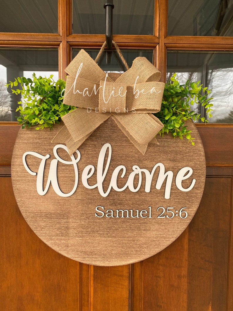 K&ouml;nnte beinhalten: Ein rundes, h&ouml;lzernes Willkommensschild mit einer Schleife aus Sackleinen und Gr&uuml;n. Das Schild tr&auml;gt die Aufschrift "Willkommen" in wei&szlig;er Schrift und "Samuel 25:6" in kleinerer wei&szlig;er Schrift.