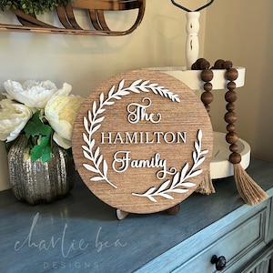 May include: A round wooden sign with a white wreath and the text "The Hamilton Family" in white lettering.