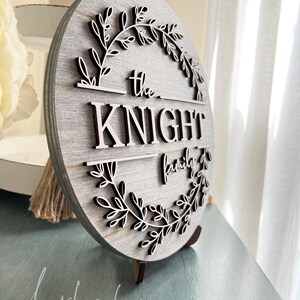 Puede incluir: Un letrero redondo de madera con un acabado de madera gris. El letrero presenta una corona de ramas frondosas que rodean el texto "the KNIGHT family".