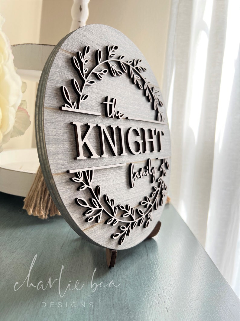 Puede incluir: Un letrero redondo de madera con un acabado de madera gris. El letrero presenta una corona de ramas frondosas que rodean el texto "the KNIGHT family".