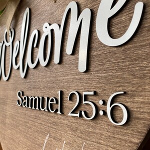 K&ouml;nnte beinhalten: Ein herzf&ouml;rmiges Willkommensschild aus Holz mit den Worten "Welcome" und "Samuel 25:6" in silbernen Lettern. Das Schild ist mit einer Schleife aus Sackleinen und gr&uuml;nem Laub verziert.