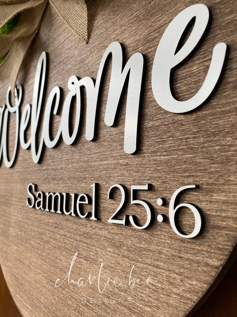 K&ouml;nnte beinhalten: Ein herzf&ouml;rmiges Willkommensschild aus Holz mit den Worten "Welcome" und "Samuel 25:6" in silbernen Lettern. Das Schild ist mit einer Schleife aus Sackleinen und gr&uuml;nem Laub verziert.