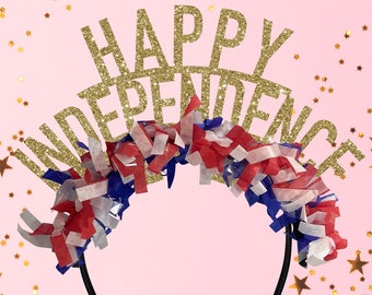 Puede incluir: Una diadema de purpurina dorada con las palabras "Happy Independence" en ella. La diadema está decorada con serpentinas rojas, blancas y azules y está rodeada de estrellas doradas brillantes sobre un fondo rosa.