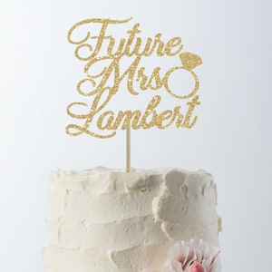 May include: Gold glitter cake topper with the text "Future Mrs. Lambert" and a diamond ring graphic. The topper is on a white cake with pink flowers.