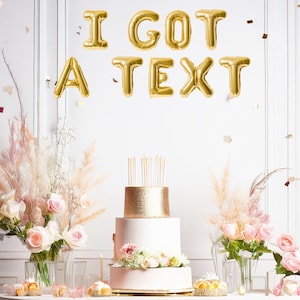May include: A two-tiered cake with gold glitter and pink frosting is on a white table with a white tablecloth. The cake is decorated with pink flowers. The background is a white wall with gold foil balloons that spell out "I GOT A TEXT".