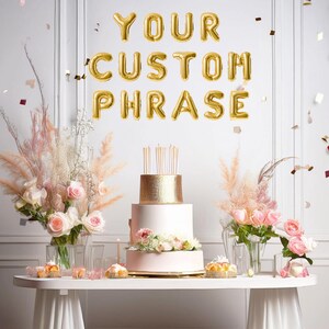 May include: A white table with a white tablecloth and gold glitter on the floor. There is a three-tiered cake with gold glitter on the bottom tier and pink frosting on the top two tiers. There are flowers on the top tier of the cake. There are two vases with pink flowers and tall dried grasses on the table. There are gold balloons spelling out "YOUR CUSTOM PHRASE" on the wall behind the table.