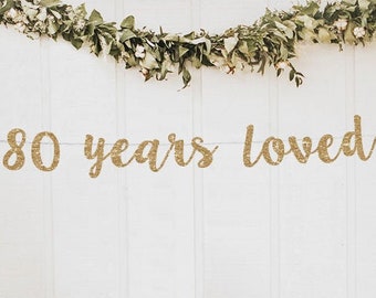 May include: Gold glitter banner with the text "80 years loved" hanging on a white wall decorated with greenery. The banner is perfect for an 80th birthday party or anniversary celebration.