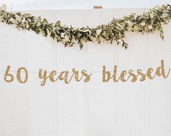 May include: Gold glitter banner with the text "60 years blessed" hanging on a white wall decorated with greenery. The banner is perfect for a 60th anniversary party.