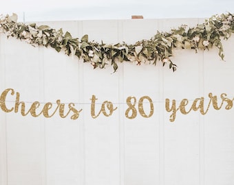 May include: Gold glitter banner with the text "Cheers to 80 years" hanging on a white wall with greenery garland.