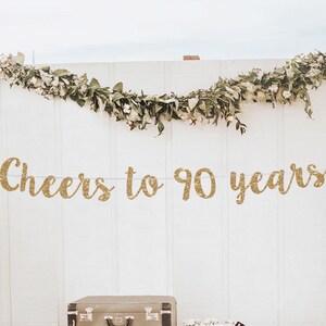 May include: Gold glitter banner with the text "Cheers to 90 years" hanging on a white wall with greenery garland. The banner is perfect for a 90th birthday party.