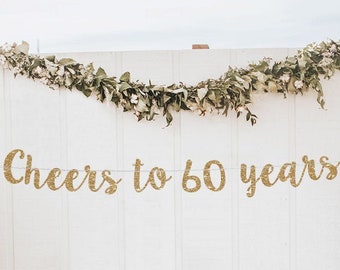 May include: Gold glitter banner with the text "Cheers to 60 years" hanging on a white wall with greenery garland above it. The banner is decorated with small gold glitter dots.