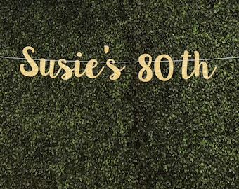 May include: Gold glitter banner with the text "Susie's 80th" hanging against a green backdrop.