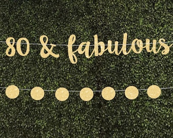 May include: A gold glitter banner that reads "80 & fabulous" is strung across the top of the image. Below the banner are six gold glitter circles. The background is a dense green hedge.