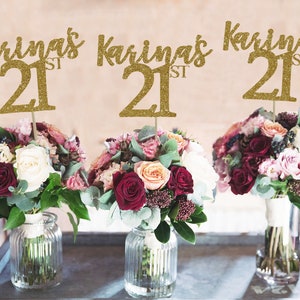 May include: Three gold glitter table centerpieces with the name "Karina's 21st" on each. The centerpieces are placed in front of three bouquets of flowers, each in a clear glass vase.