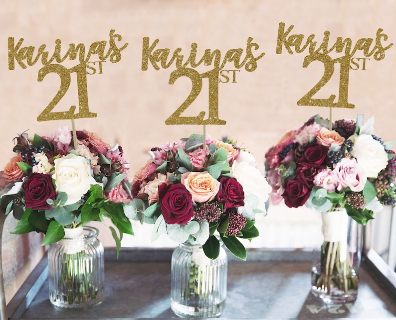 May include: Three gold glitter table centerpieces with the name "Karina's 21st" on each. The centerpieces are placed in front of three bouquets of flowers, each in a clear glass vase.