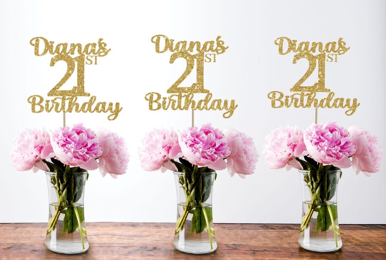 May include: Three gold glitter table centerpieces with the text "Diana's 21st Birthday" on each. The centerpieces are displayed in clear glass vases with pink peonies.