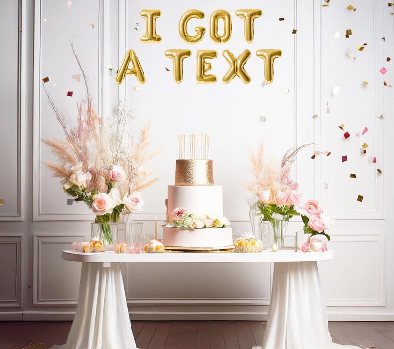 May include: A two-tiered cake with gold glitter and pink frosting is on a white table with a white tablecloth. The cake is decorated with pink flowers. The background is a white wall with gold foil balloons that spell out "I GOT A TEXT".