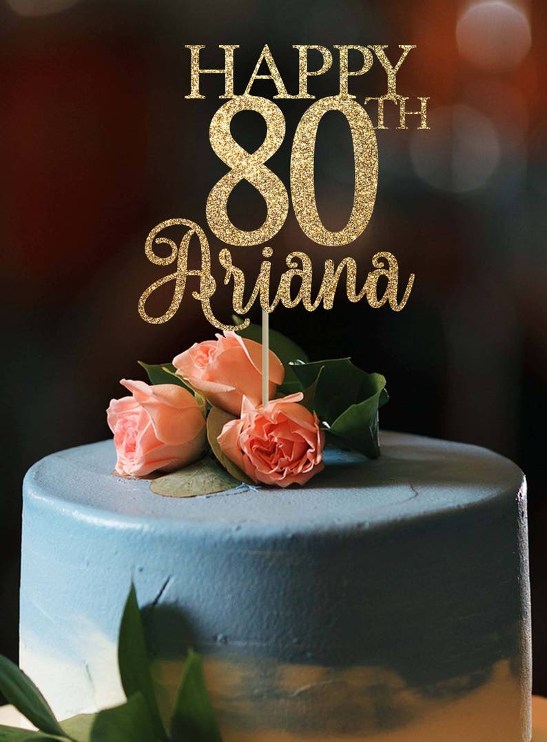 Puede incluir: Topper de pastel de purpurina dorada con el texto "Happy 80th Ariana" en un pastel degradado azul y blanco con rosas rosadas.