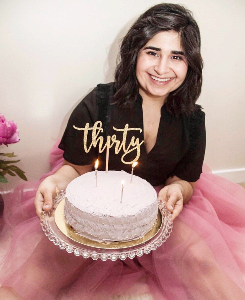 K&ouml;nnte beinhalten: Eine Frau in einem schwarzen Oberteil und einem rosa T&uuml;llrock h&auml;lt einen lila gefrosteten Kuchen mit Kerzen und einem goldenen Glitzer-Kuchentopper "drei&szlig;ig".