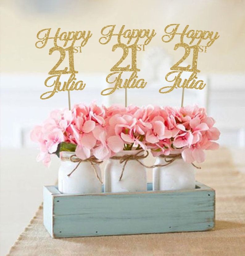 May include: Three gold glitter Happy 21st Julia centerpieces with pink flowers in white mason jars in a light blue wooden box.