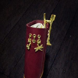 May include: A burgundy leather journal with gold detailing. The journal has gold dragonfly and flower charms, and a gold pen holder. A gold elastic band wraps around the journal. The journal is open, revealing white pages.