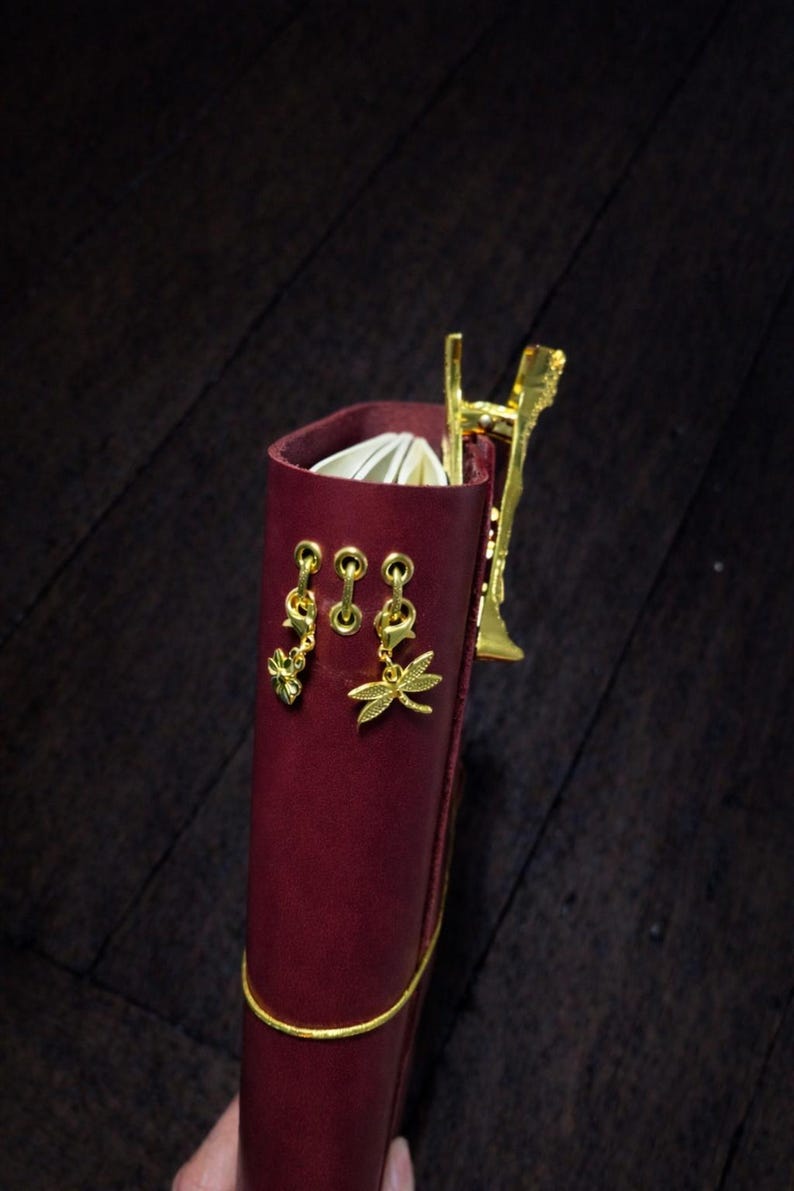 May include: A burgundy leather journal with gold detailing. The journal has gold dragonfly and flower charms, and a gold pen holder. A gold elastic band wraps around the journal. The journal is open, revealing white pages.