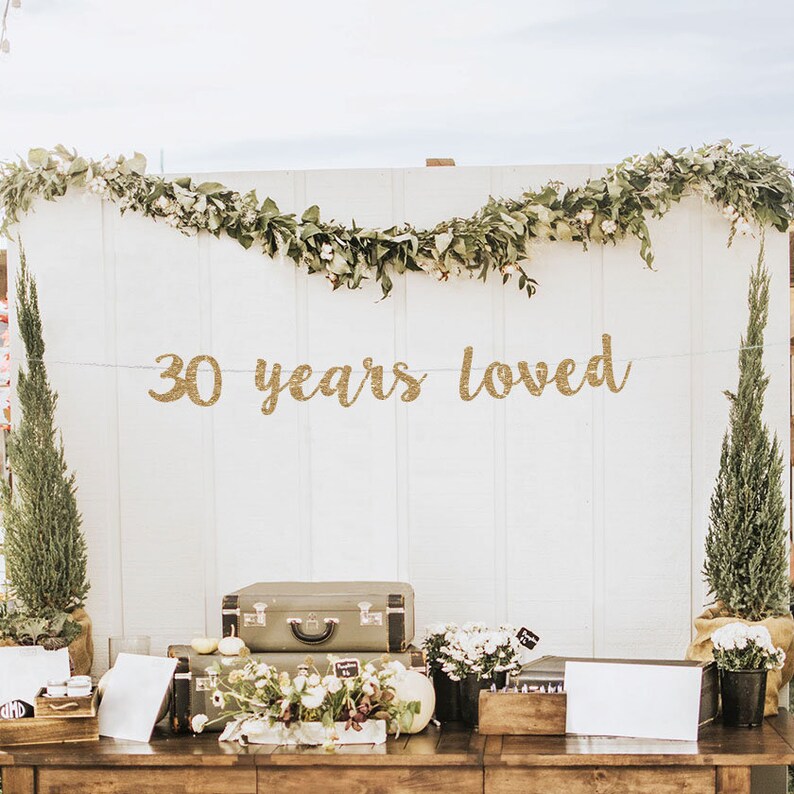 May include: Gold glitter banner with the text "30 years loved" hanging on a white wall decorated with greenery. The banner is perfect for a 30th anniversary party.