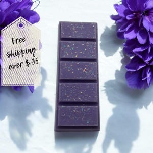 May include: A rectangular, dark purple wax melt bar with five sections, speckled with colorful glitter. The bar is centered on a white surface, flanked by vibrant purple flowers. The image includes a tag with the text "Free Shipping over $35".