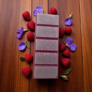 Può includere: Una barra rettangolare di cera profumata con cinque sezioni in una tonalità lilla chiaro, cosparsa di glitter, su una base rosa. Lamponi rossi freschi e fiori viola sono sparsi intorno alla barra su una superficie di legno, creando un tema naturale.
