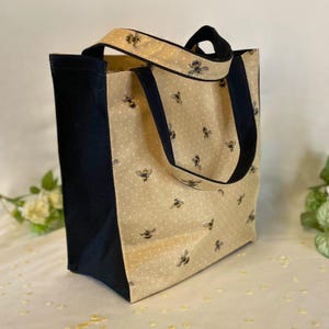 Peut inclure: Un sac fourre-tout rectangulaire avec un fond beige, des pois blancs et des illustrations d'abeilles. Le sac a des c&ocirc;t&eacute;s et des poign&eacute;es bleu marine. Les poign&eacute;es sont bord&eacute;es du motif abeille et pois.